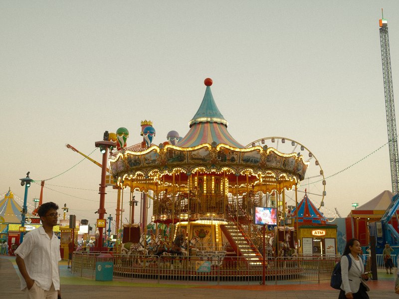 A brightly lit carousel at a fairground during twilight, with people nearby enjoying the atmosphere.