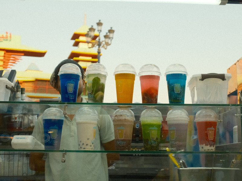 A vendor behind a glass display of colorful drinks in cups, with reflections of a street scene.