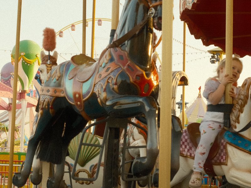 A child riding a carousel horse at a carnival during golden hour, with colorful balloons in the background.