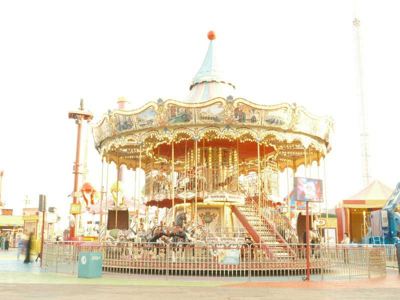 A brightly lit carousel with ornate decorations and a blue-topped canopy, surrounded by amusement park rides.