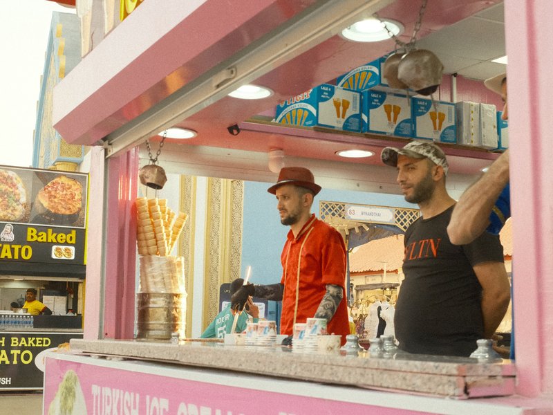 Two men in a colorful ice cream stall, one wearing a red shirt and the other in a black t-shirt.