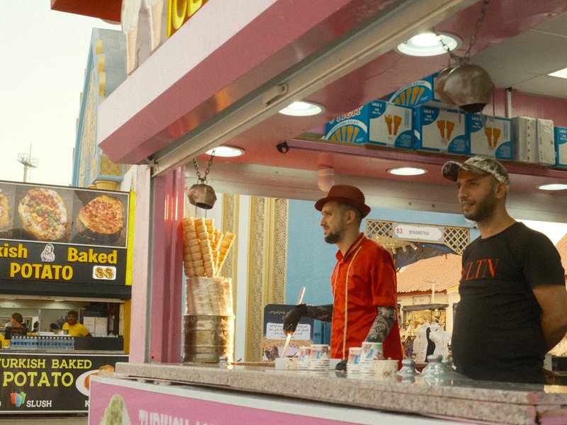 Two men in a food stall with colorful signage, one wearing a red jacket and the other in a black t-shirt.