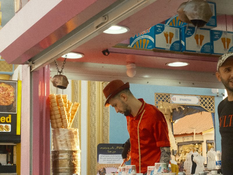 Two men working at an ice cream stand with colorful decor and ice cream cones displayed.