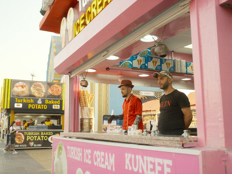 Two men at an ice cream stand with a pink exterior, colorful signage, and food stalls in the background.