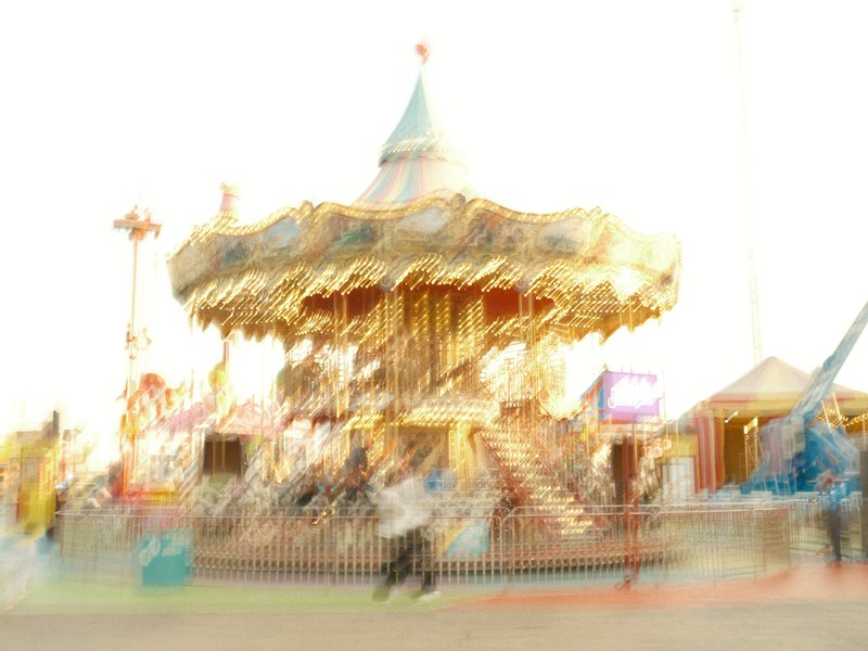 A blurred image of a carousel at a fair, with bright lights and a festive atmosphere.