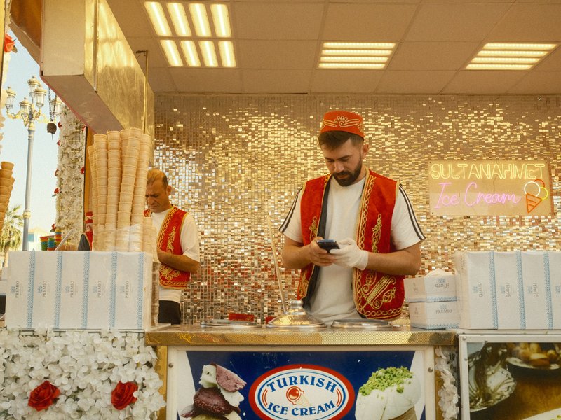 A man in a red vest checks his phone at an ice cream stand decorated with flowers and sparkles.