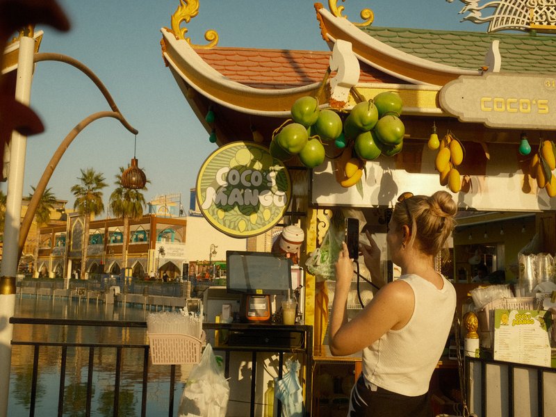 A woman stands at a colorful outdoor fruit stand with tropical fruits and a pond in the background.