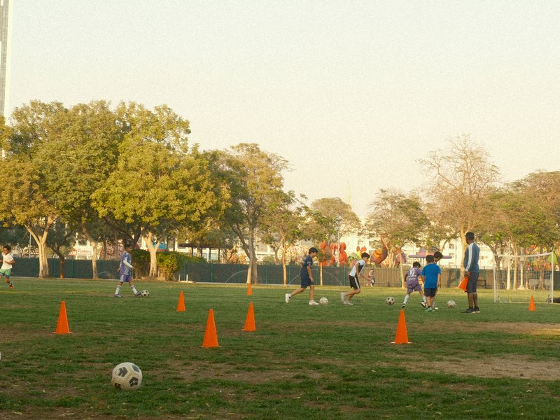 Children practicing soccer on a grassy field with orange cones and trees in the background.