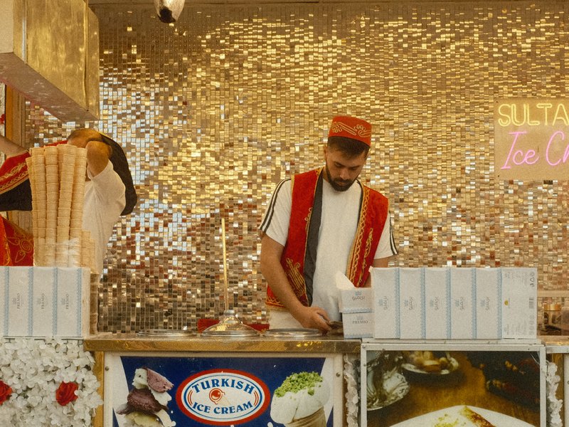 A vendor in traditional attire prepares food at a vibrant stall with a sparkling backdrop.