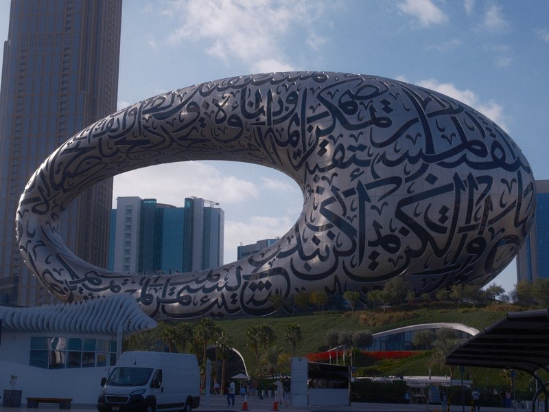 A modern architectural structure resembling an oval shape with Arabic calligraphy, surrounded by buildings.