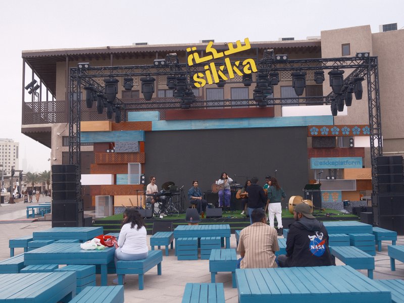 Outdoor stage with musicians performing, colorful seating area, and modern architecture in the background.