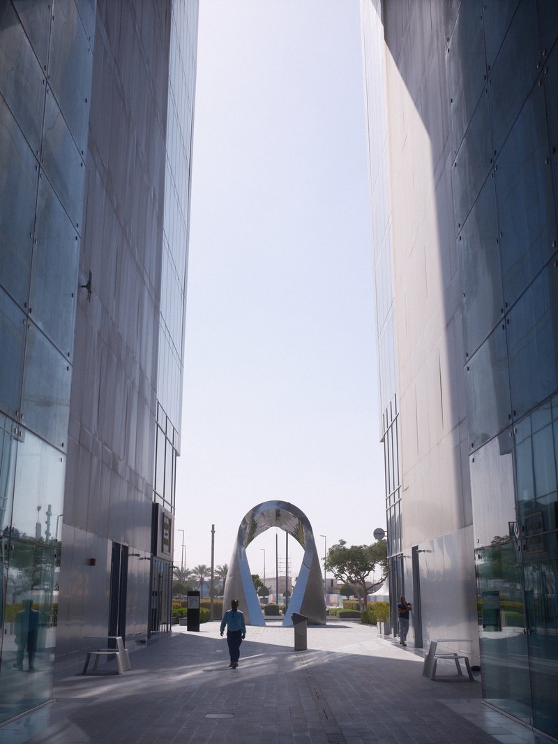 A person walking through a modern architectural passageway with reflective glass walls and a sculpture.