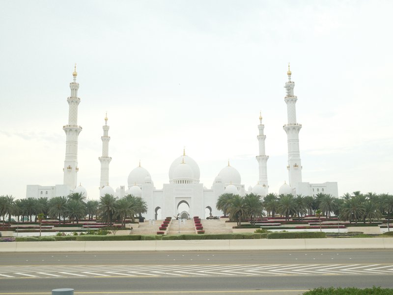 A grand white mosque with multiple domes and minarets, surrounded by greenery and a cloudy sky.