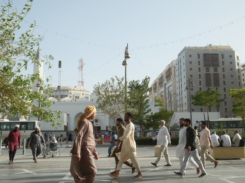 People walking in a busy urban area with buildings and trees under a clear sky.