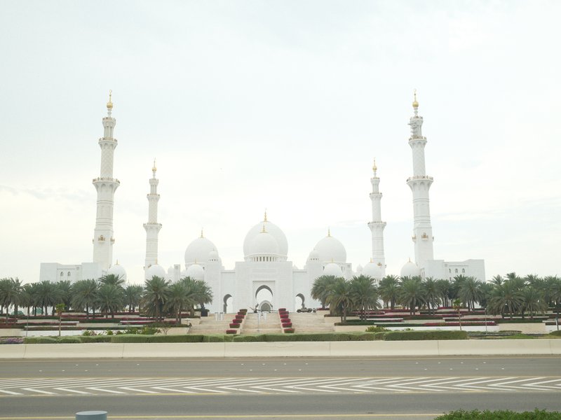 A grand white mosque with tall minarets, surrounded by greenery and a wide road in front.