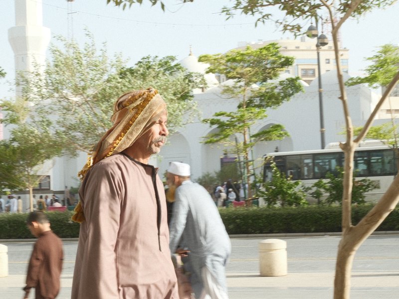 A man in traditional attire walking in an urban setting with trees and a bus in the background.