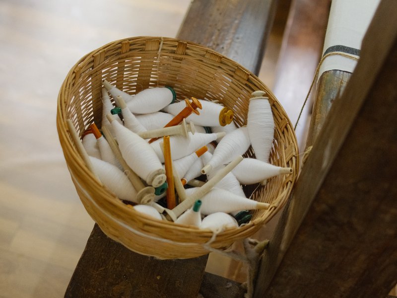 A woven basket filled with white cotton spools and wooden spindles resting on a wooden surface.