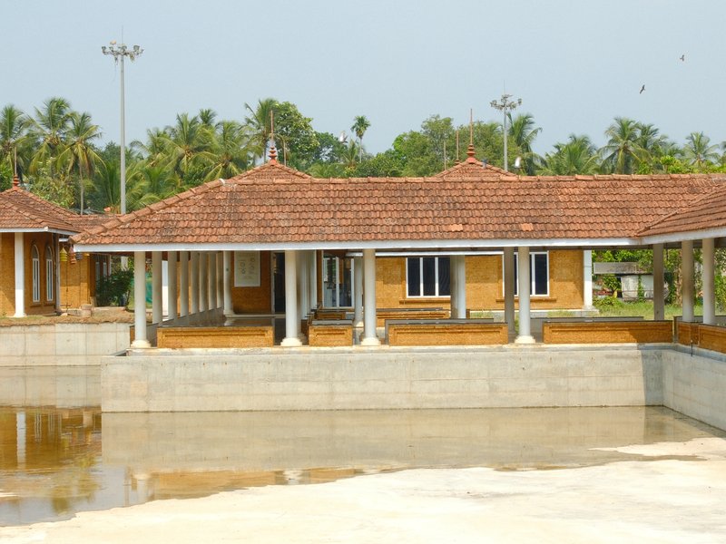 A traditional building with a tiled roof beside a reflective water body, surrounded by greenery.