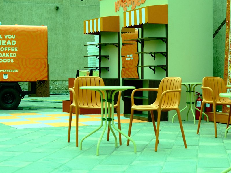 A vibrant outdoor café scene with orange chairs and a green table, featuring a coffee truck in the background.
