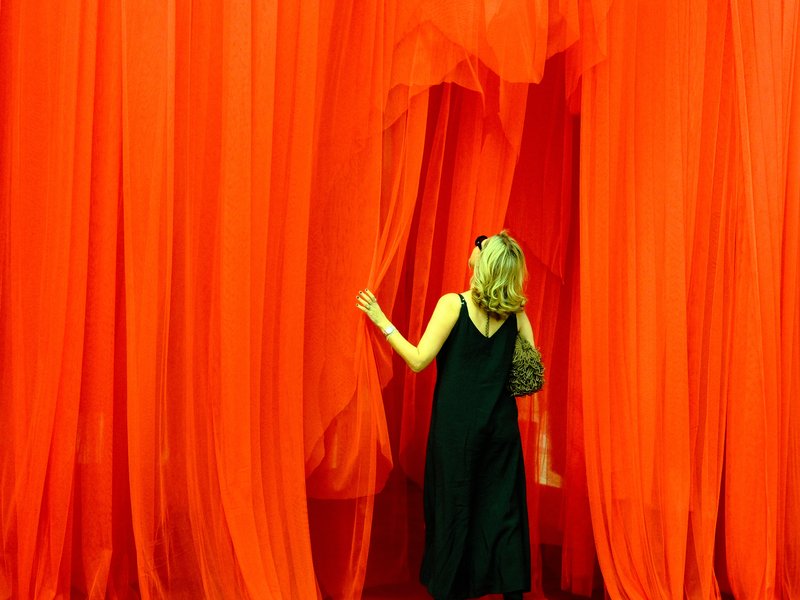 A woman in a black dress stands behind flowing red curtains, partially revealing her figure.