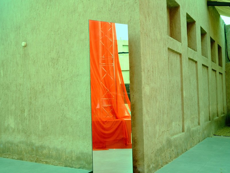 A tall mirror reflecting an orange drape against a textured green wall.
