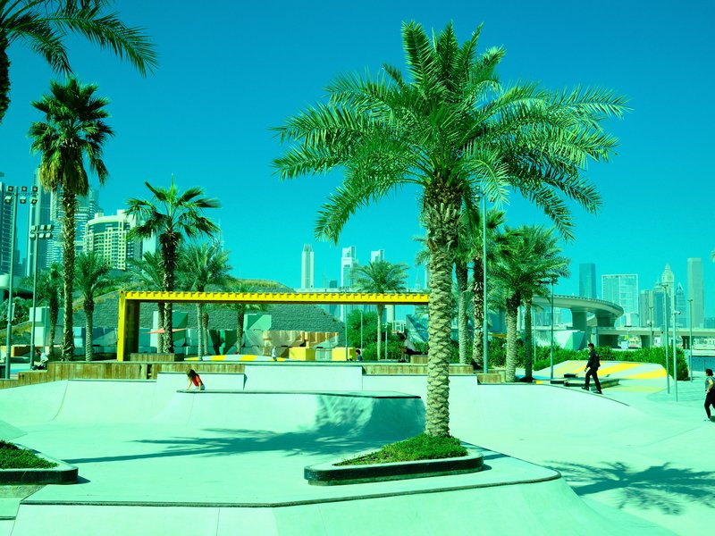 A vibrant urban park scene featuring palm trees, a yellow structure, and people enjoying the space.