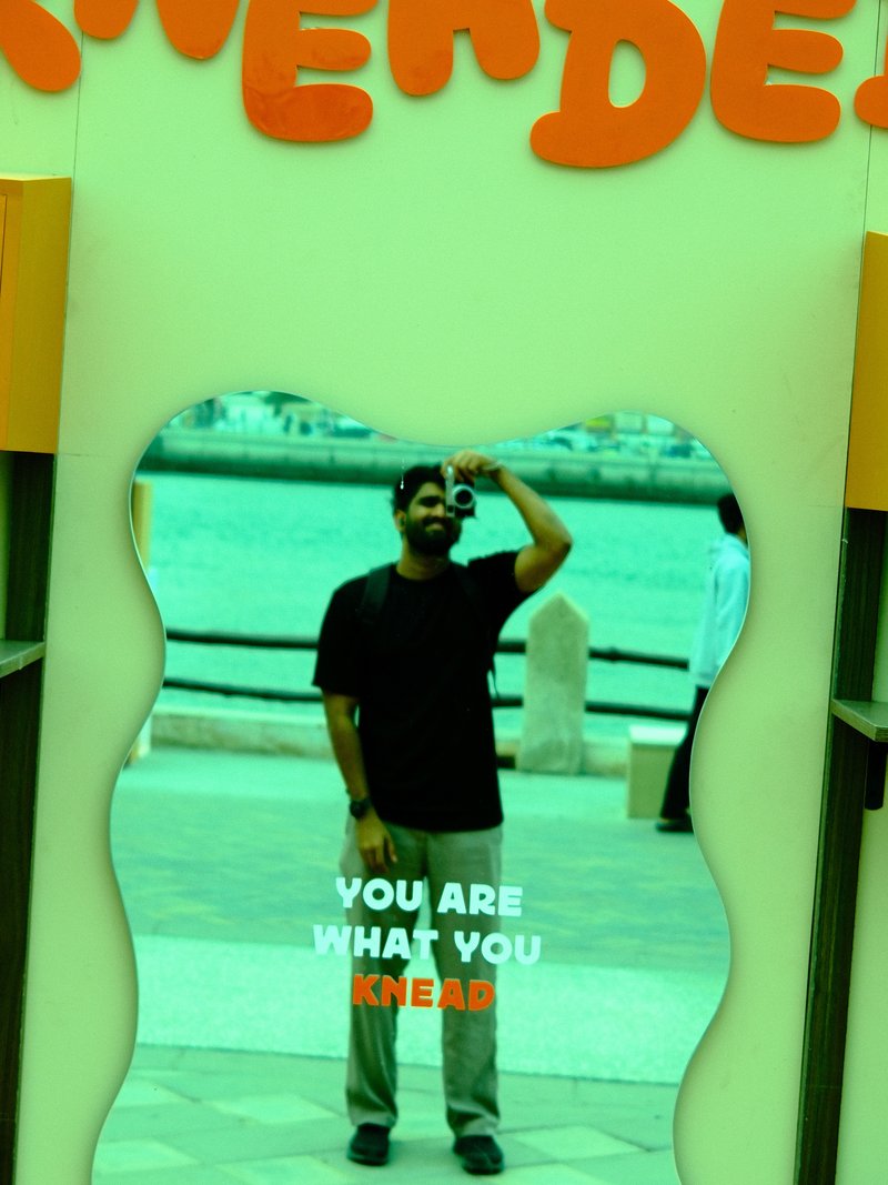A person standing in front of a colorful mirror with a playful message, by a waterfront.