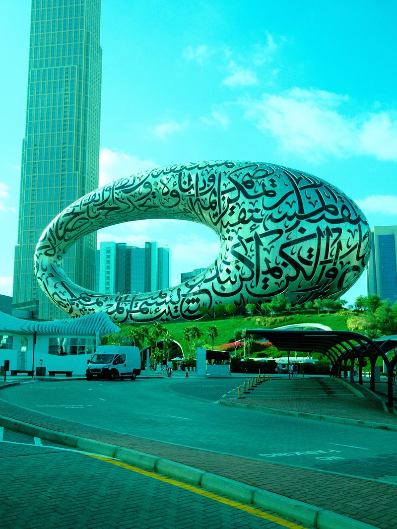 A large, modern sculpture with Arabic calligraphy, set against a city skyline.