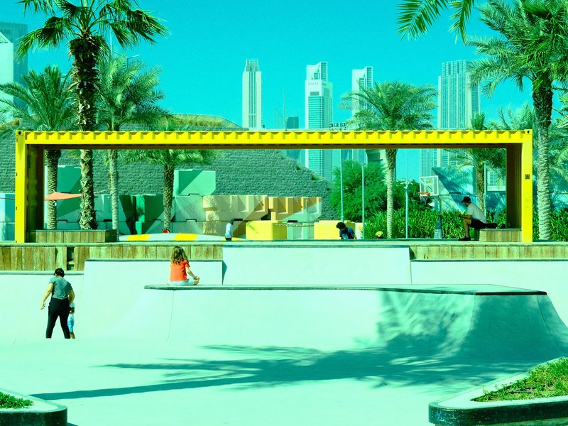 A skate park with a yellow frame structure, palm trees, and city skyline in the background.
