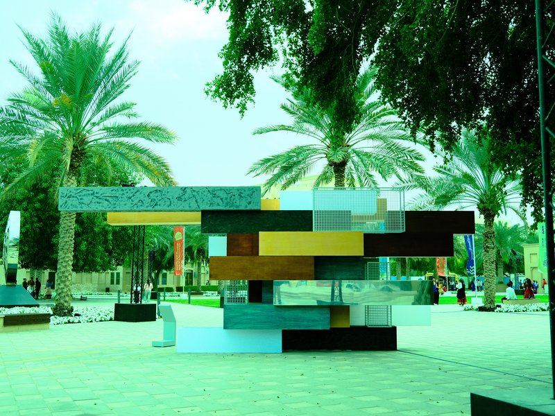 A modern sculpture made of colorful geometric shapes in a park surrounded by palm trees.