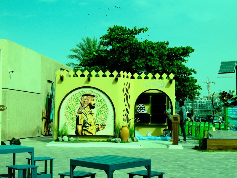 A colorful outdoor scene featuring a mural of a man in traditional attire, surrounded by greenery and furniture.