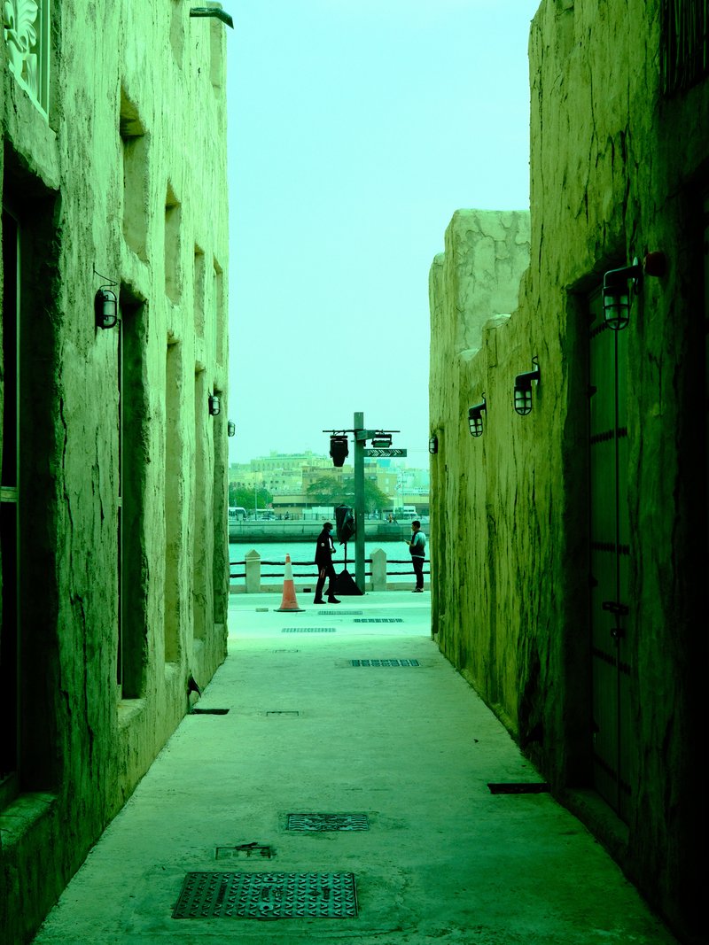 Narrow alleyway between textured walls leading to a waterfront view with pedestrians in the distance.