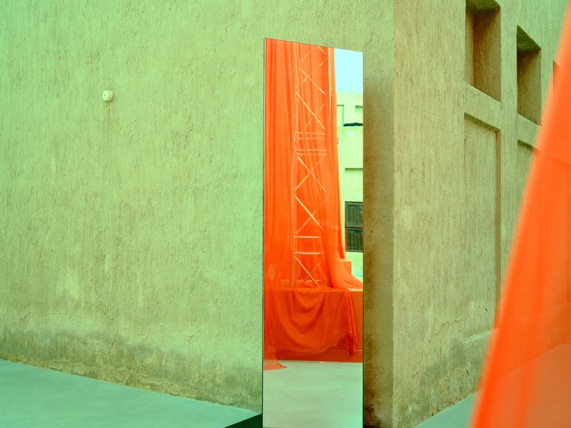 A reflective mirror stands against a textured wall, draped in vibrant orange fabric.