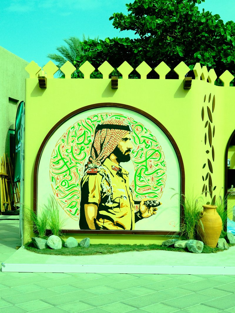 A vibrant mural of a man in traditional attire against a bright green wall, surrounded by plants.