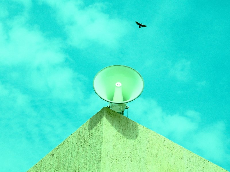 A green megaphone mounted on a building corner under a bright turquoise sky with a bird flying above.