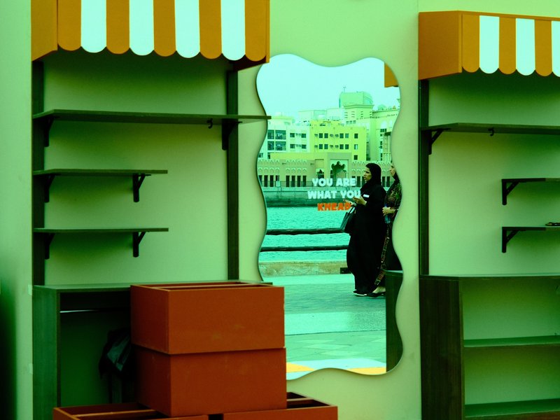 A mirror reflects a figure in traditional attire near colorful market stalls by the water.