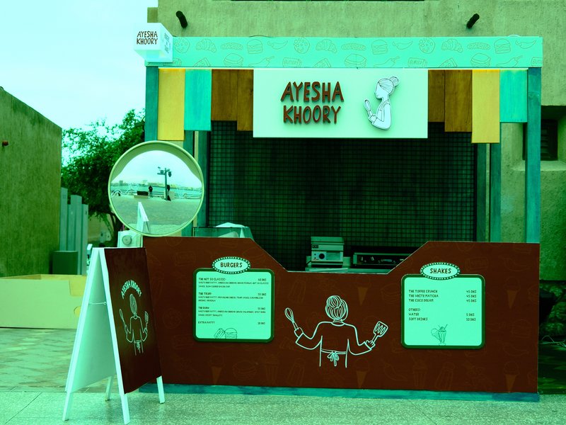 A colorful food stall named 'Ayesha Khory' with a mirror and menu displayed.