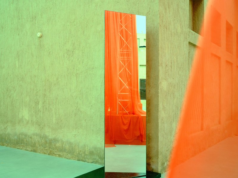 A tall mirror reflecting an orange curtain against a textured wall, with vibrant green tones in the scene.