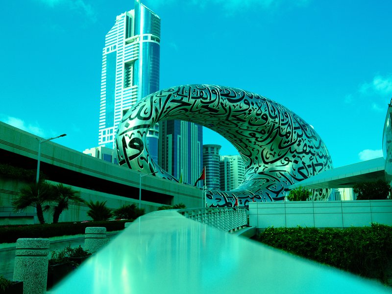 A modern architectural structure with intricate patterns, surrounded by tall buildings under a bright blue sky.