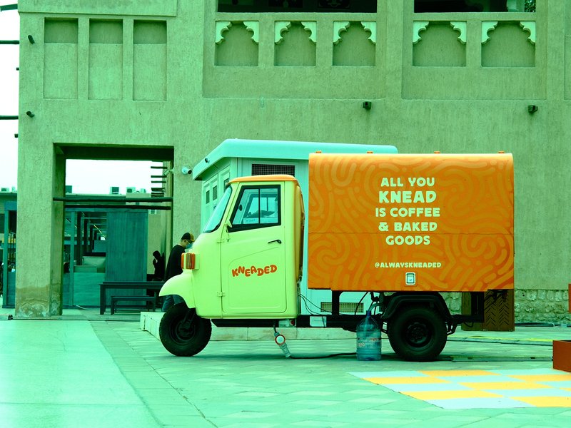 A colorful food truck with a bright orange sign offering coffee and baked goods, set in an urban plaza.