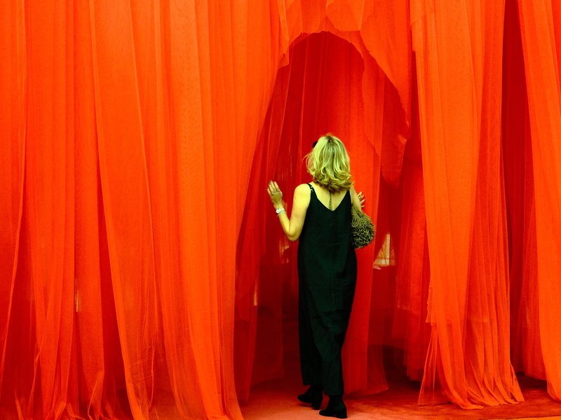 A woman in a black dress walks through flowing red fabric curtains in a vibrant, artistic setting.