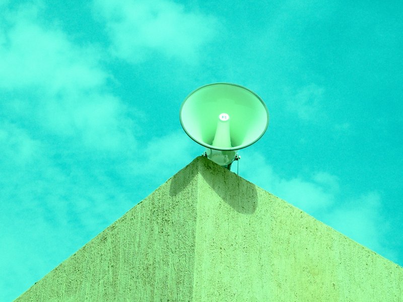 A green megaphone mounted on a corner of a building against a bright turquoise sky.