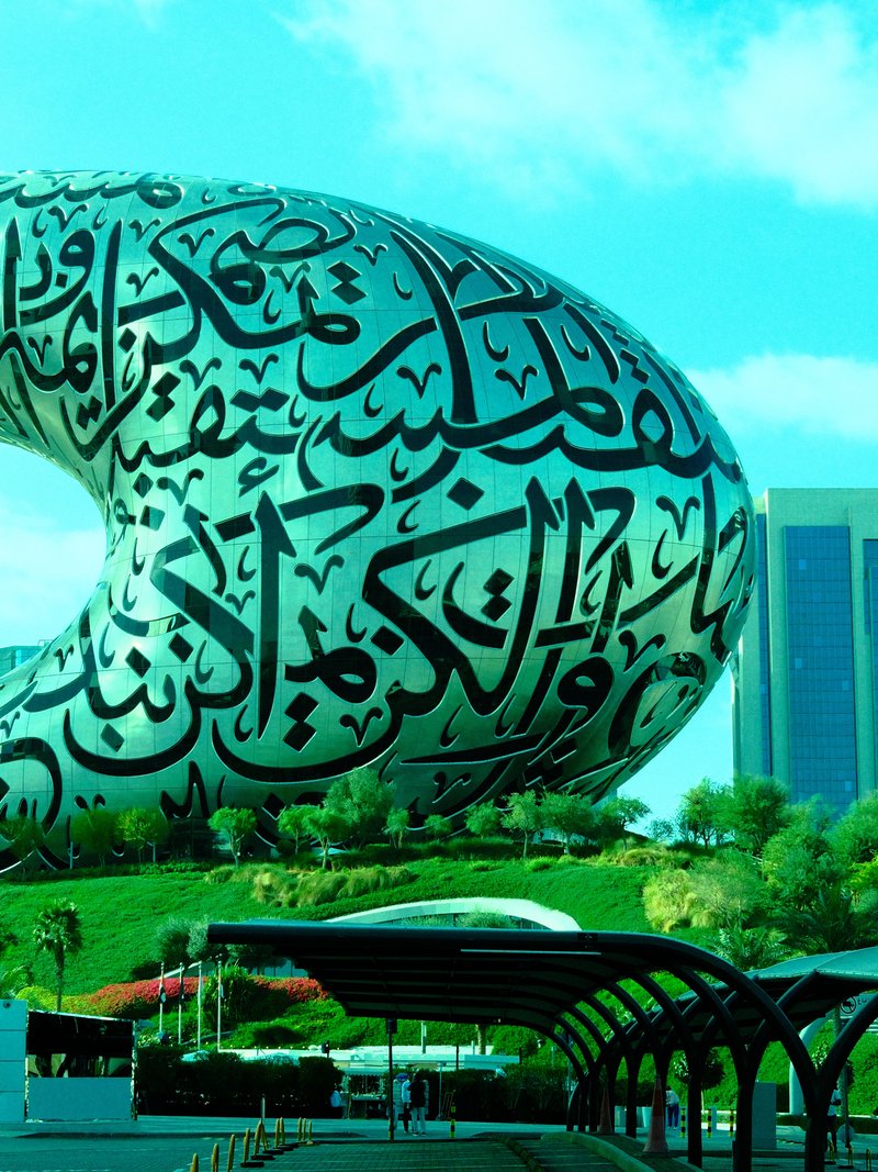 A large, modern sculpture covered in Arabic calligraphy, set against a blue sky and green landscape.