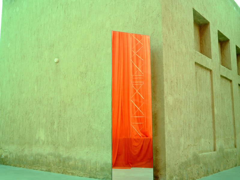 A bright orange fabric draped against a textured beige wall with geometric cutouts.