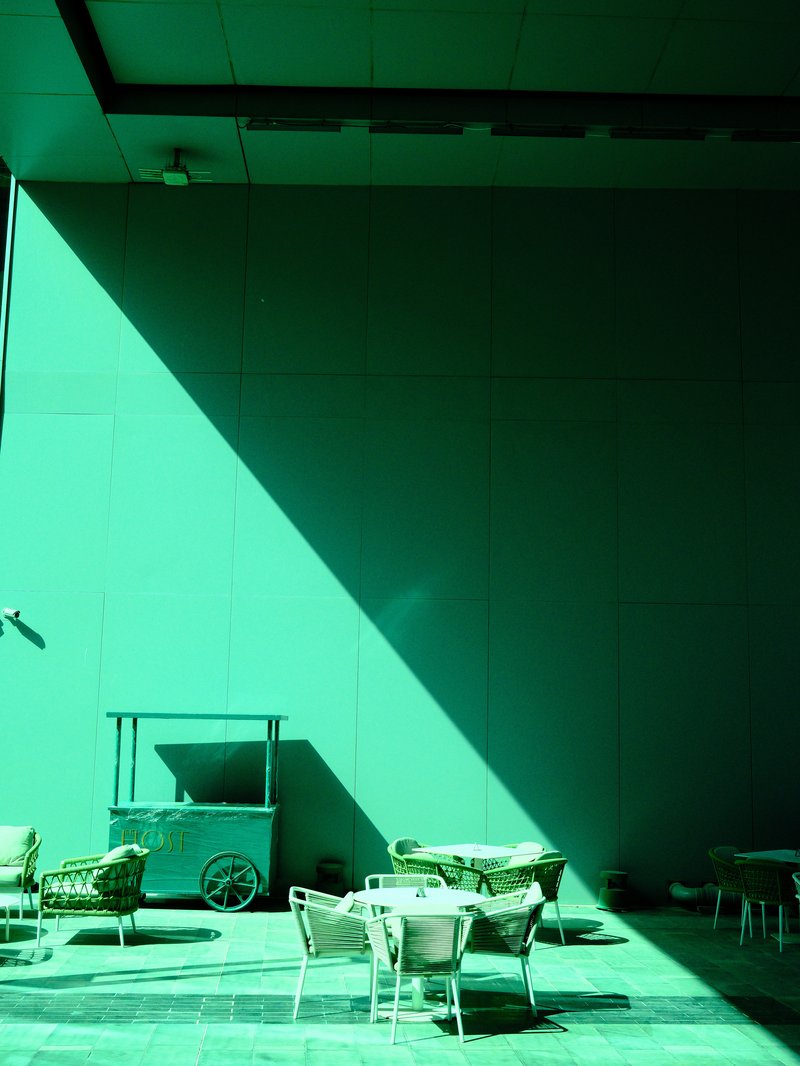 A green wall with a long shadow, featuring empty tables and chairs in a modern outdoor setting.
