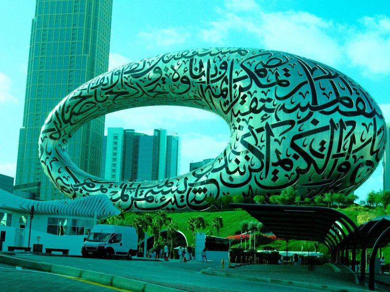 A large, abstract sculpture covered in Arabic calligraphy, set against a modern city skyline.