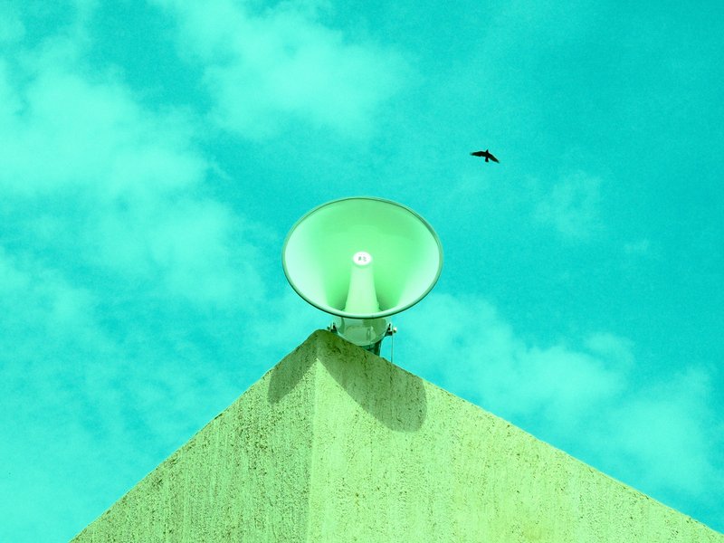 A green megaphone atop a textured wall under a bright blue sky with scattered clouds.
