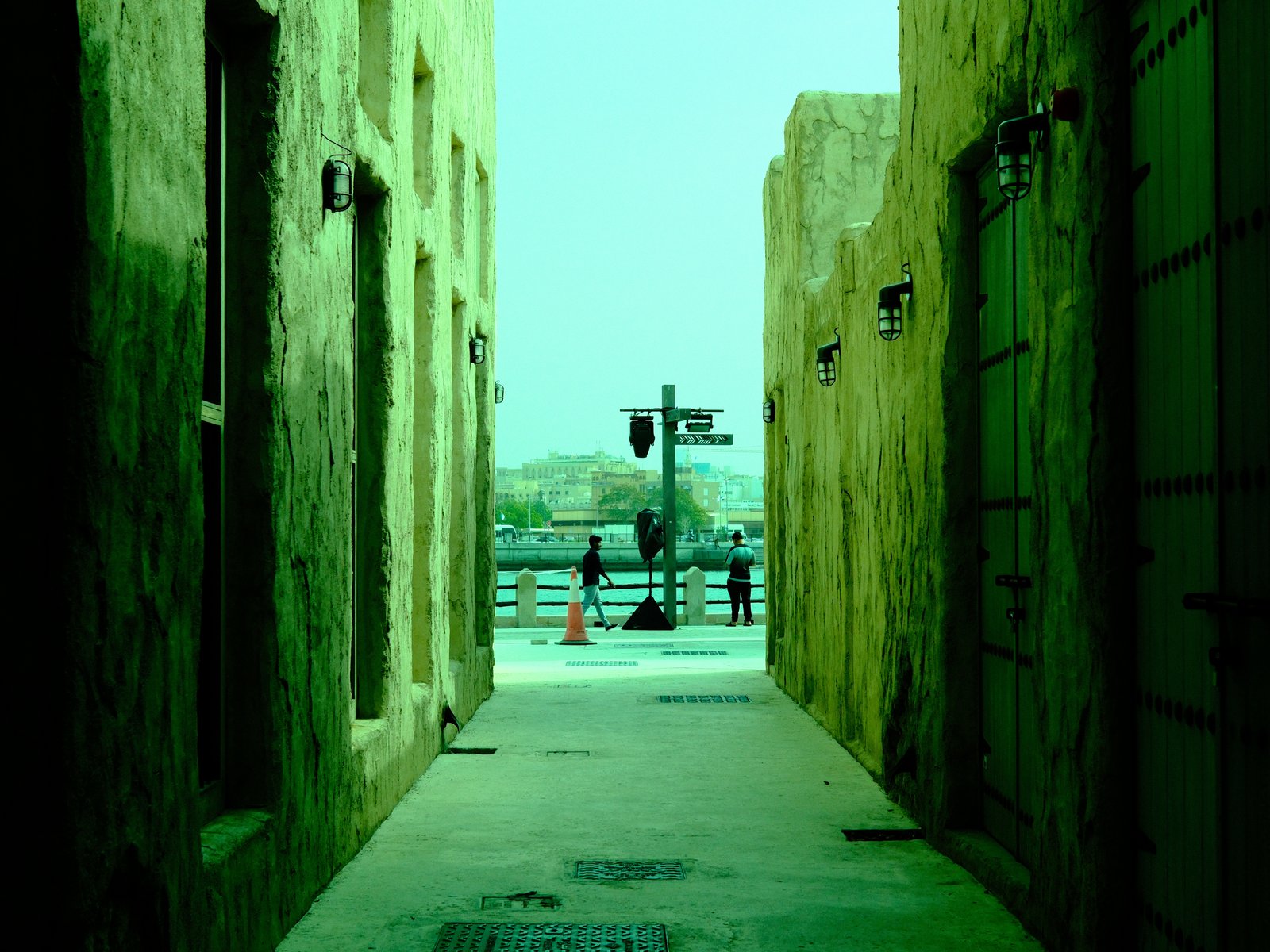 Narrow alleyway with greenish tint leading to a waterfront view, people in the distance.