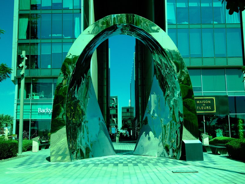A reflective arch sculpture in an urban setting, surrounded by modern buildings and greenery.