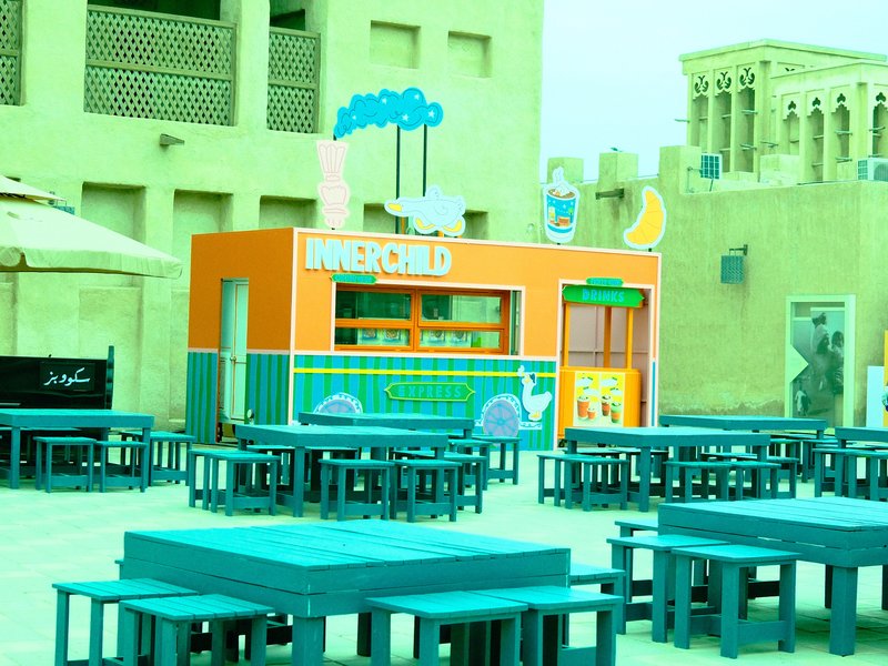 Colorful food stall with blue tables in an outdoor setting, surrounded by beige buildings.